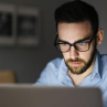 A man wearing glasses sitting at a desk looking at his laptop computer.