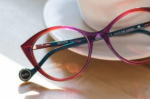 A pair of pink and blue eyeglasses are folded on a table leaning against a coffee mug.