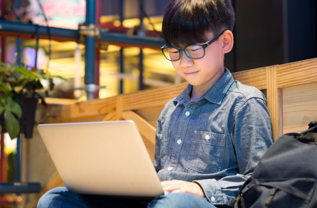 A happy young boy is wearing blue light glasses and doing his homework on his laptop computer while sitting outside.