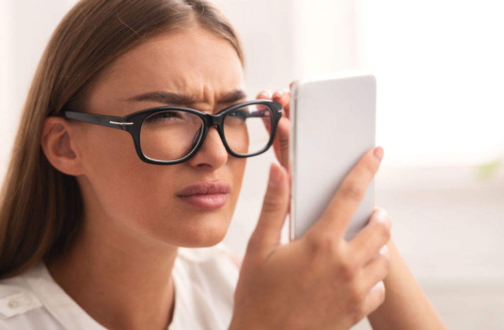 A woman wearing glasses, squinting, and holding her phone close to her face to see it better.