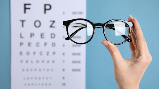 A hand holds up a pair of black round eyeglasses in front of a blurred eye chart on a blue background, representing a changing vision prescription.