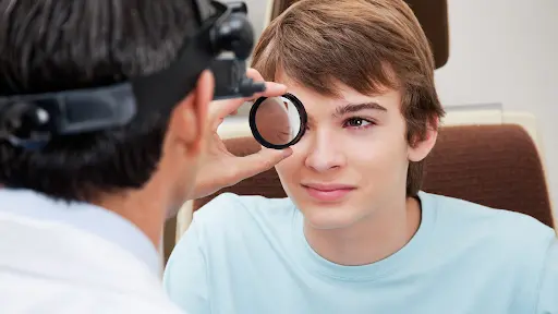 An eye doctor wearing a headband ophthalmoscope holds a handheld lens up to a young man's eye during a dilated eye exam.